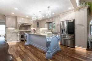 Elegant traditional kitchen featuring custom cream cabinetry with raised panel doors, an ornate wood range hood with carved detailing, and a statement island with decorative blue-gray corbels. Granite countertops and crystal chandeliers add a luxurious finishing touch alongside warm natural wood flooring.